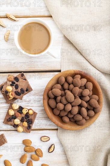 Almond in chocolate dragees in wooden plate and a cup of coffee on white wooden background and