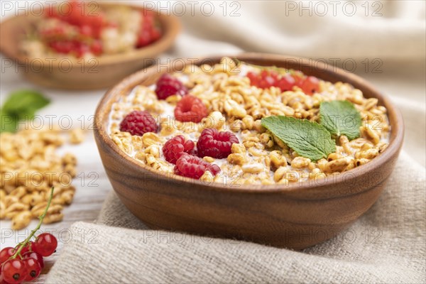 Wheat flakes porridge with milk, raspberry and currant in wooden bowl on white wooden background