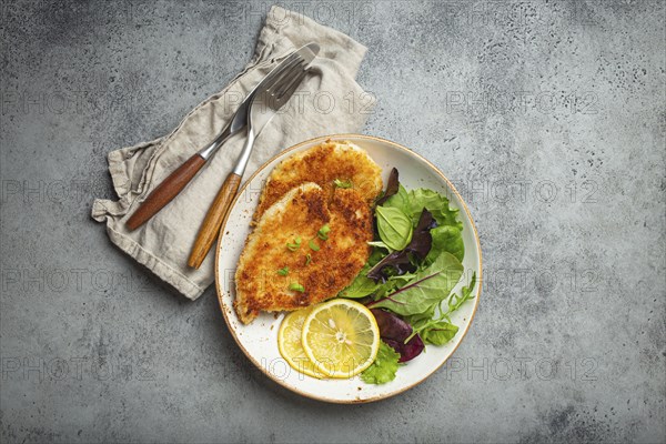 Crispy panko breaded fried chicken fillet with green salad and lemon on plate with cutlery on gray