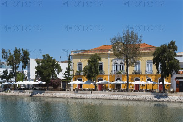 Yellow building with terrace area by the river, surrounded by trees and a restaurant in the