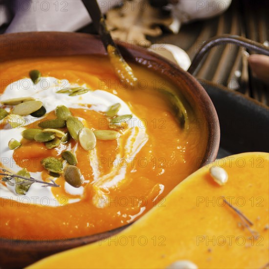 Clay dish with homemade rustic pumpkin soup with seeds on wooden table with bread and greens aside