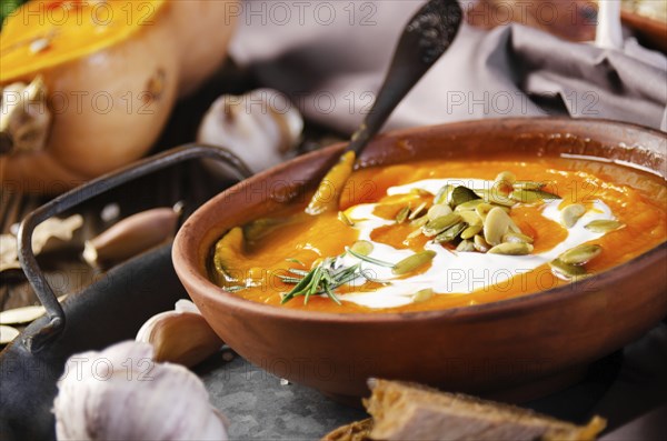 Clay dish with homemade rustic pumpkin soup with seeds on wooden table with bread and greens aside