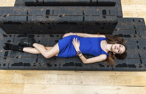 Portrait of an 18 year old white woman, in a blue short dress, lying on a grey couch, view from