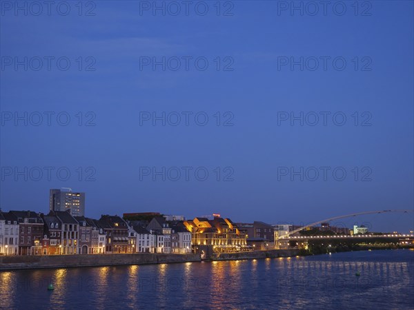 Riverbank with illuminated houses and a bridge during the blue hour, maastricht, the netherlands
