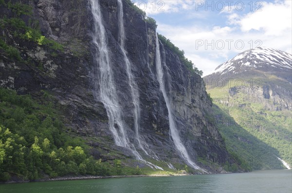 High waterfalls flow down a steep cliff face, surrounded by green forests and mountains under a