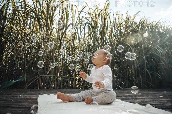 A baby on a blanket surrounded by sunlight, playing with soap bubbles