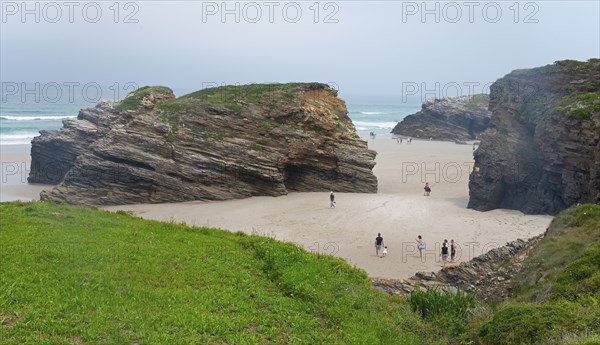 People walking on the beach between rocky cliffs and green grass under a cloudy sky, Praia das
