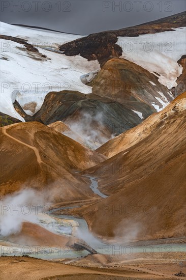 Bridge over steaming stream between colourful rhyolite mountains with snowfields, Hveradalir