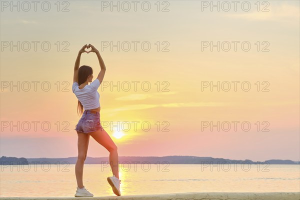 Woman raised her hands up and folded her palms in shape of heart against backdrop of sunset