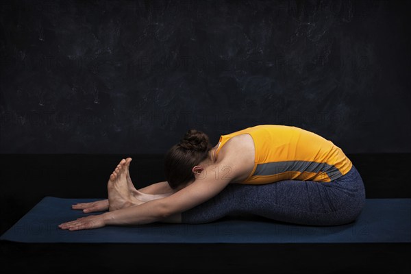 Woman doing Ashtanga Vinyasa Yoga back bending asana Paschimottanasana, seated forward bend on dark