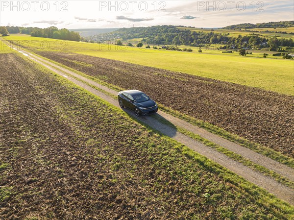 A black car drives along a country lane, surrounded by vast fields under a cloudy sky, Carsharing,