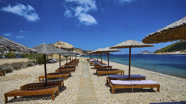 A beach with a wooden walkway and deckchairs under thatched umbrellas, the view of the clear water