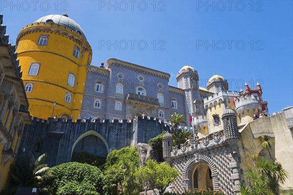 Castle with grey and yellow towers against a blue sky, surrounded by plants, Palácio Nacional da