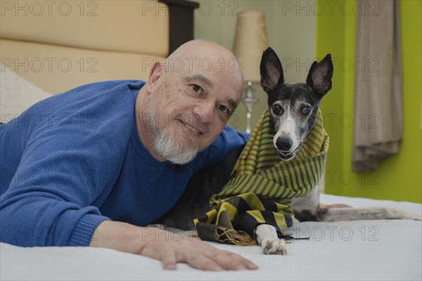 Mature man posing and smiling while looking at the camera with his greyhound pet on the bed