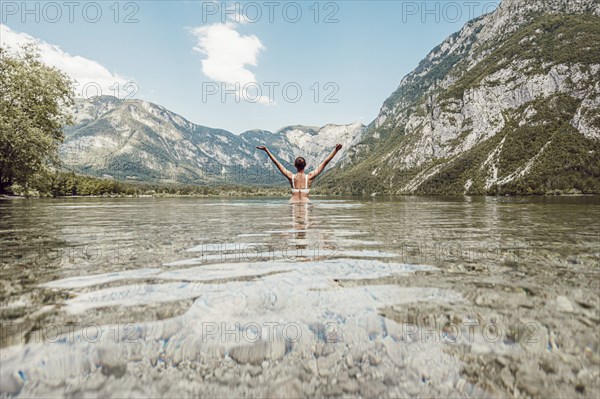 Woman with arms outstretched to the sky on both sides in a clear lake, surrounded by mountains and