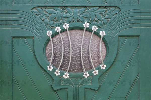 Close-up of a green door with elaborate decorations and a small oval glass window with a metal