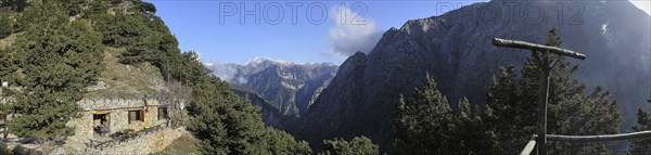 Mountain hut nestled in a wooded mountain landscape with views of snow-capped peaks under a blue