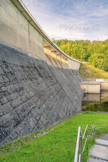 Concrete dam surrounded by green grass and lush forest in the background in sunny weather,