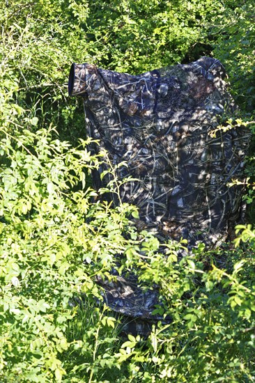 Nature photographer in camouflage at work, camouflage net in a dense bush surrounded by green