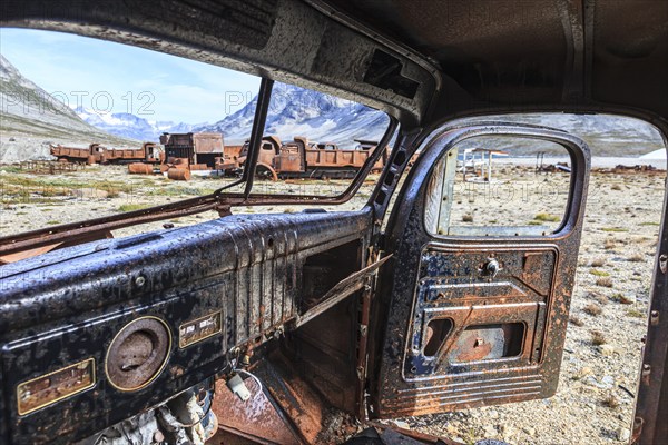 Interior view of a rusty car in front of steep mountains, remains of a US airbase from the Second
