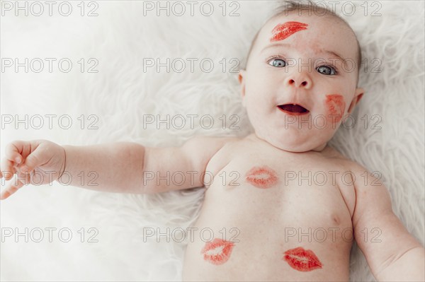A smiling baby with red lipstick kisses on her face and body lies on a white blanket and looks into