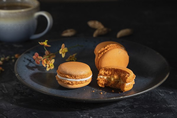 Orange macarons or macaroons cakes with cup of coffee on a black concrete background. Hard light,