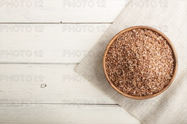 Wooden bowl with unpolished brown rice on a white wooden background and linen textile. Top view,