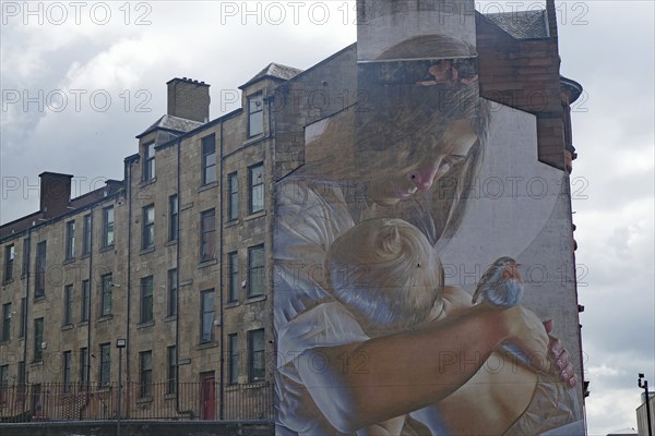 A mural shows a woman holding a baby and a bird in her arms. A building can be seen in the