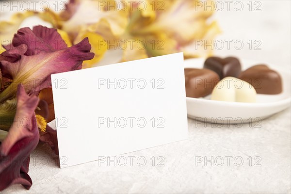 White business card with chocolate candies and iris flowers on gray concrete background. side view,