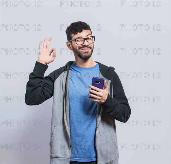 Pleasant person holding cellphone and winking, smiling boy with cellphone making ok sign, concept