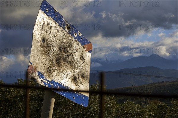 A rusty, tattered road sign in front of a mountainous landscape under a cloudy sky, Lefka Ori,
