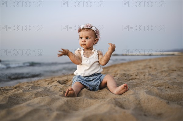 A thoughtful baby in summery clothes and flowers in her hair sits on the beach at sunset