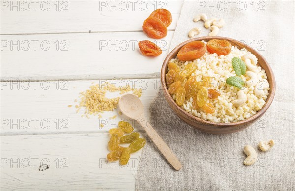 Bulgur porridge with dried apricots, raisins and cashew in wooden bowl on a white wooden background