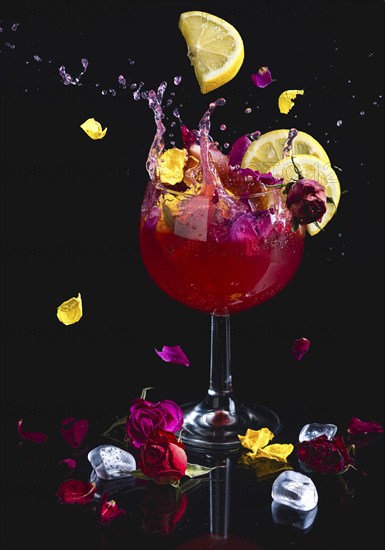 A colourful splashing cocktail with lemon slices, surrounded by flowers, reflection, dark