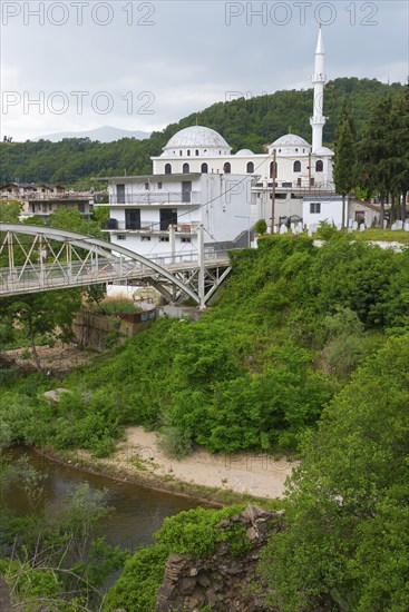 A mosque with a white dome and minaret next to a bridge over a river, surrounded by greenery,