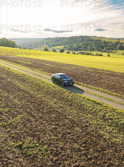 Black car driving on a gravelled country lane through a wide, rural landscape under a cloudy sky,