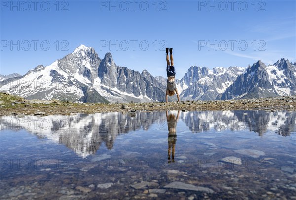Young man doing a handstand in front of a mountain landscape, reflection in a small mountain lake,