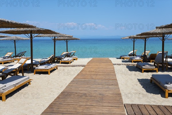 Wooden walkway leads to a sunlit beach with deckchairs and parasols by the clear sea, Lago Seaside