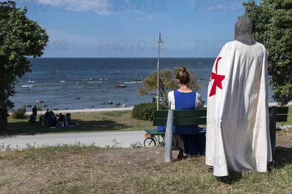 A knight with a red cross on a white coat and armour stands behind a woman sitting on a bench with