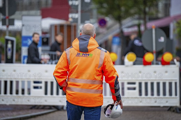 A worker in an orange safety waistcoat with a helmet stands in front of a barrier on the road,