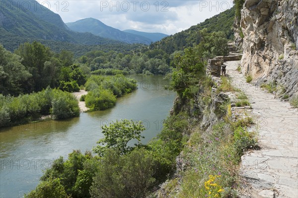 A rocky hiking trail along the bank of a river with surrounding mountain views, River Nestos or