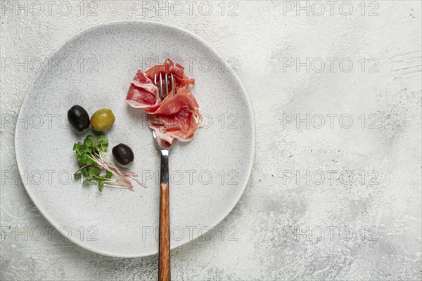 Prosciutto on a fork, top view, olives and micro-greens on a plate, light background, no people