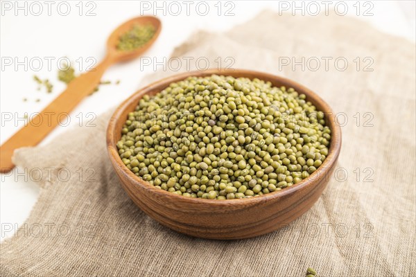 Wooden bowl with raw green mung bean and wooden spoon on a white background and linen textile. Side