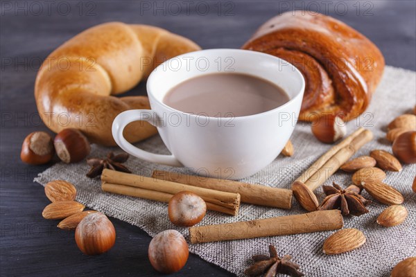 A cup of hot chocolate with nuts, buns and spices on linen napkin and black wooden background.