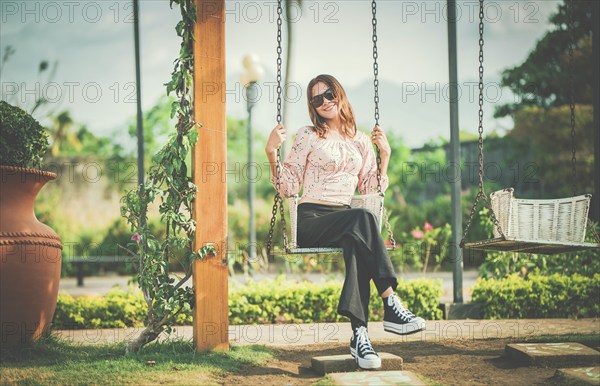 Happy young woman sitting on a swing in a nature park. Portrait of a smiling girl sitting on a