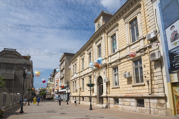 A lively city street with historic architecture and balloon decorations on a sunny day, pedestrian