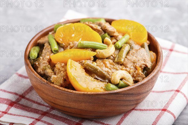 Fried pork with peaches, cashew and green beans in a wooden bowl on a gray concrete background.