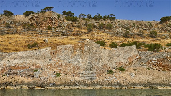 Old stone wall on rocky cliff with dry vegetation and calm sea water in the foreground, Venetian