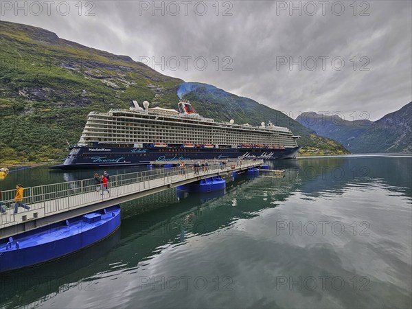 A large cruise ship, Mein Schiff 6, lies in the fjord, surrounded by green mountains under a cloudy