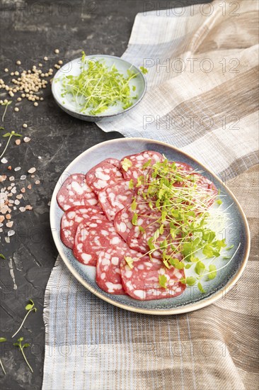 Slices of smoked cervelat salami sausage with spinach microgreen, salt and pepper on black concrete